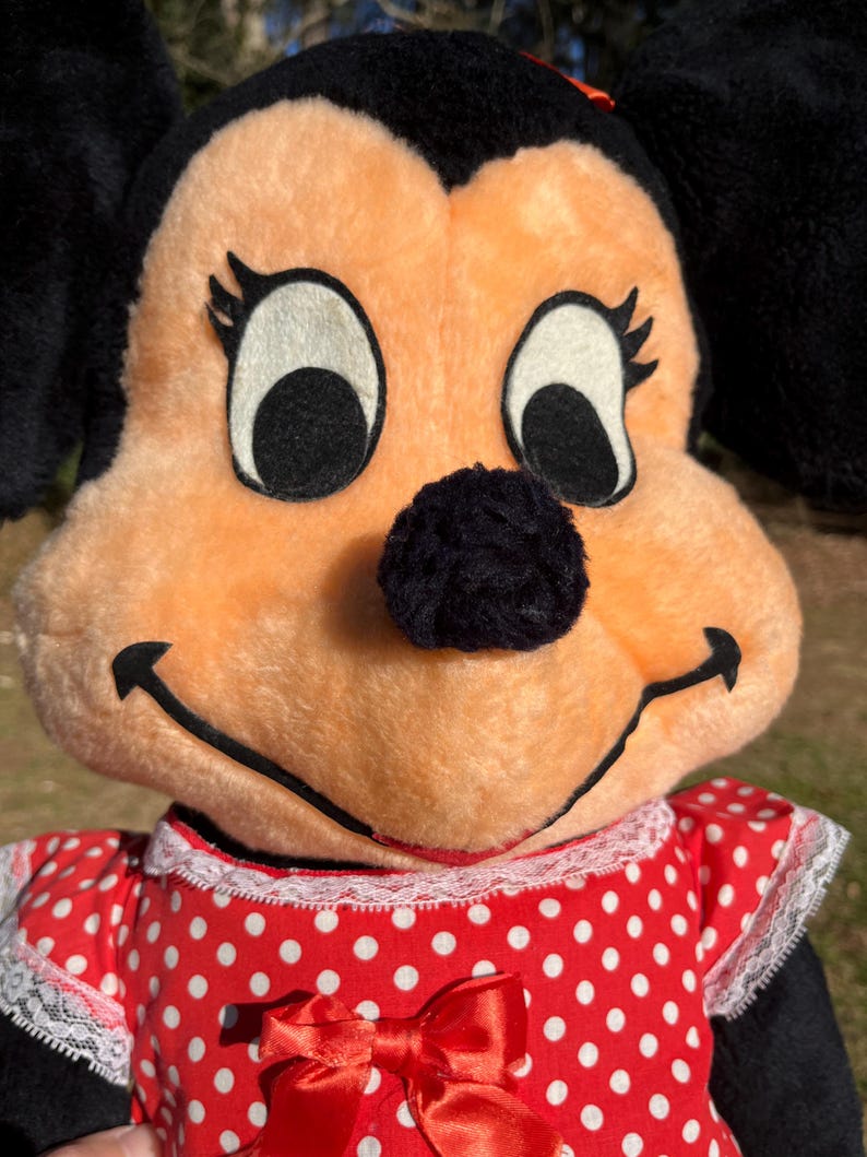 Op de afbeelding: Close-up van een Minnie Mouse pluche pop. De pop heeft een perzikkleurig gezicht, zwarte oren en een zwarte neus. Ze draagt een rode jurk met witte stippen en een rode strik. De pop heeft grote, zwarte ogen en een glimlachende uitdrukking.