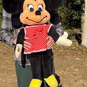 Op de afbeelding: Een grote Minnie Mouse pluche pop. De pop heeft zwarte oren, een perzikkleurig gezicht en een rode jurk met witte stippen. De schoenen van de pop zijn geel en ze heeft zwarte benen en armen. De pop wordt door een persoon vastgehouden.