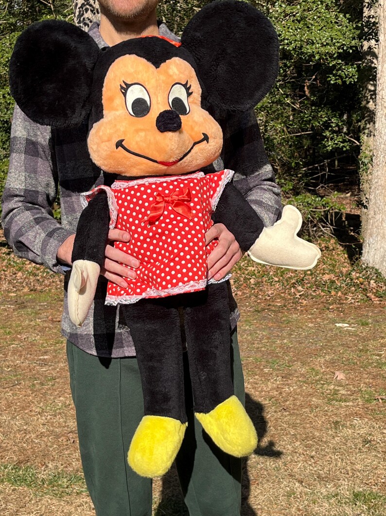 Op de afbeelding: Een grote Minnie Mouse pluche pop. De pop heeft zwarte oren, een perzikkleurig gezicht en een rode jurk met witte stippen. De schoenen van de pop zijn geel en ze heeft zwarte benen en armen. De pop wordt door een persoon vastgehouden.