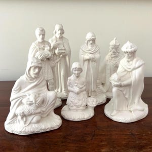 May include: A collection of white ceramic nativity figurines, including figures of the three wise men, a shepherd, and a young girl. The figurines are arranged on a dark wooden surface.