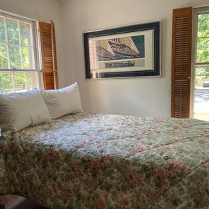May include: A bed with a dark wood spindle frame, two white pillows, and a quilted coverlet featuring a green and pink floral pattern. Above the bed hangs a framed print of a sailboat, flanked by two windows with wooden shutters on each side.