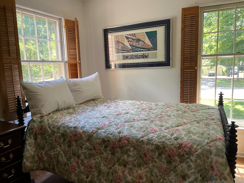 May include: A bed with a dark wood spindle frame, two white pillows, and a quilted coverlet featuring a green and pink floral pattern. Above the bed hangs a framed print of a sailboat, flanked by two windows with wooden shutters on each side.