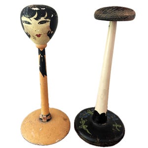 May include: Two vintage hat forms, one with a painted face and the other with a black and white floral design. Both are made of wood and have a painted finish.