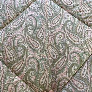 May include: A quilted fabric featuring a repeating paisley pattern in shades of sage green, soft pink, and cream. The textile displays intricate teardrop-shaped motifs with detailed floral accents, stitched together in a grid-like diamond pattern.