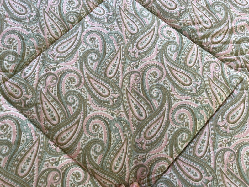 May include: A quilted fabric featuring a repeating paisley pattern in shades of sage green, soft pink, and cream. The textile displays intricate teardrop-shaped motifs with detailed floral accents, stitched together in a grid-like diamond pattern.