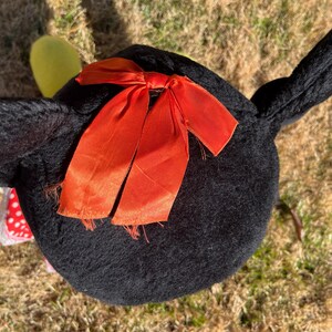 Op de afbeelding: Een zwart pluche speelgoed met een feloranje strik. Het speelgoed heeft lange, puntige oren en een ronde body. Een kleine, gele vorm is zichtbaar bij &eacute;&eacute;n oor. Het speelgoed ligt op droog gras.