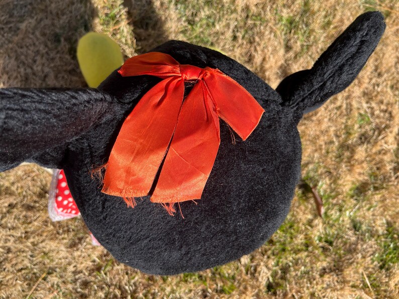 Op de afbeelding: Een zwart pluche speelgoed met een feloranje strik. Het speelgoed heeft lange, puntige oren en een ronde body. Een kleine, gele vorm is zichtbaar bij &eacute;&eacute;n oor. Het speelgoed ligt op droog gras.