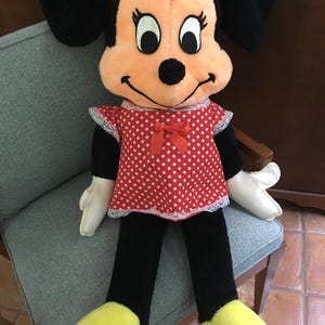 Op de afbeelding: Een Minnie Mouse pluche pop, zittend op een stoel. De pop heeft een rode jurk met witte stippen en witte kanten rand, een zwarte broek, gele schoenen en witte handschoenen. Minnie's gezicht is perzik van kleur met grote zwarte ogen en zwarte oren.