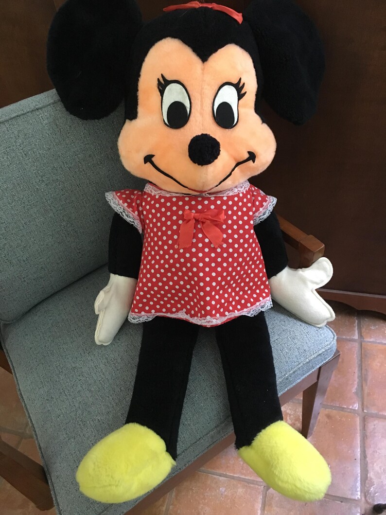 Op de afbeelding: Een Minnie Mouse pluche pop, zittend op een stoel. De pop heeft een rode jurk met witte stippen en witte kanten rand, een zwarte broek, gele schoenen en witte handschoenen. Minnie's gezicht is perzik van kleur met grote zwarte ogen en zwarte oren.