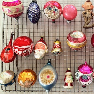 May include: A collection of vintage Christmas ornaments in various shapes and colors. Includes a red guitar, Santa Claus, bells, and round ornaments with glitter and painted designs. The ornaments are displayed on a metal grid.