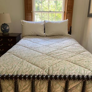 May include: A bed featuring a green and white paisley patterned quilt with a diamond-stitched texture. The bed has a dark brown wooden spindle headboard and footboard, accompanied by two white pillows and a dark wood nightstand with a lamp.