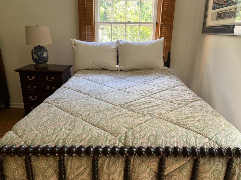 May include: A bed featuring a green and white paisley patterned quilt with a diamond-stitched texture. The bed has a dark brown wooden spindle headboard and footboard, accompanied by two white pillows and a dark wood nightstand with a lamp.
