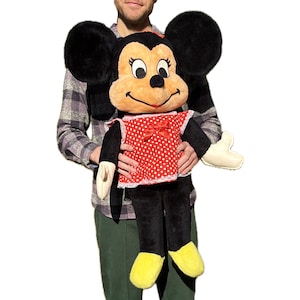 Op de afbeelding: Een grote Minnie Mouse pluche pop. De pop heeft zwarte oren van bont, een bruin gezicht en een rode jurk met witte stippen en een strik. De armen en benen van de pop zijn zwart en ze heeft gele schoenen. De pop wordt door een persoon vastgehouden.