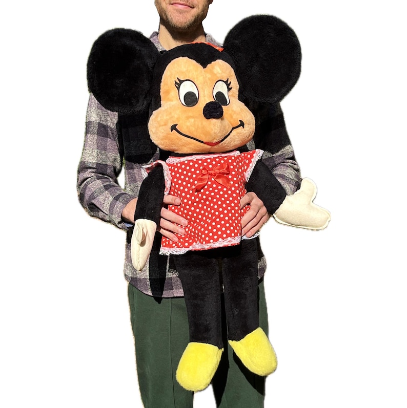 Op de afbeelding: Een grote Minnie Mouse pluche pop. De pop heeft zwarte oren van bont, een bruin gezicht en een rode jurk met witte stippen en een strik. De armen en benen van de pop zijn zwart en ze heeft gele schoenen. De pop wordt door een persoon vastgehouden.