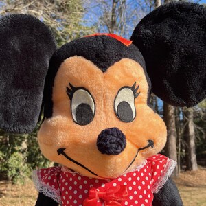 Op de afbeelding: Een pluche Minnie Mouse pop met grote zwarte oren, een perzikkleurig gezicht en een rood-witte polkadot jurk. De pop heeft een rode strik op haar hoofd en een zwarte neus. De achtergrond is wazig met bomen en zonlicht.