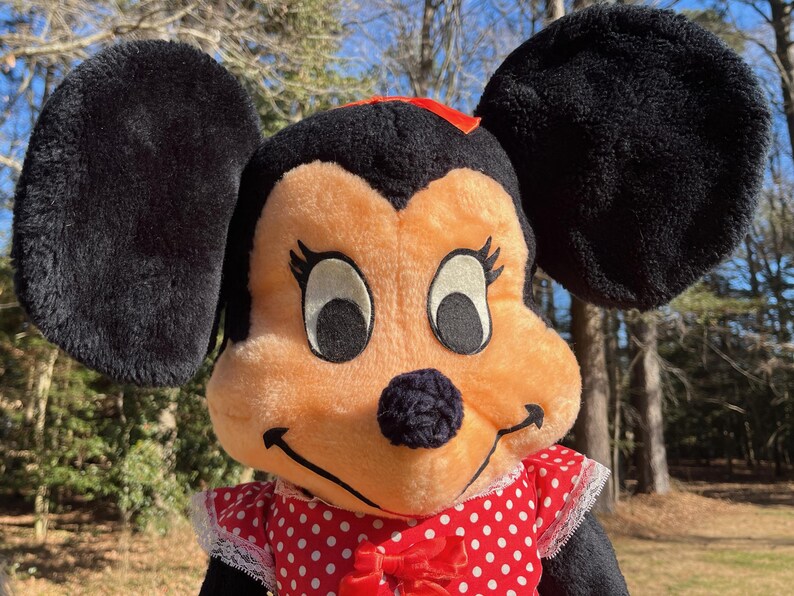 Op de afbeelding: Een pluche Minnie Mouse pop met grote zwarte oren, een perzikkleurig gezicht en een rood-witte polkadot jurk. De pop heeft een rode strik op haar hoofd en een zwarte neus. De achtergrond is wazig met bomen en zonlicht.
