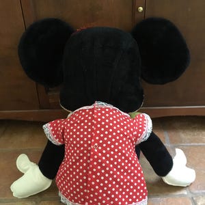 Op de afbeelding: Een Minnie Mouse knuffel, van achteren gezien, met grote zwarte oren en een rode jurk met witte stippen. De jurk heeft witte kanten randen en een kleine knoopsluiting. De armen en handen van de pop zijn zwart en wit.