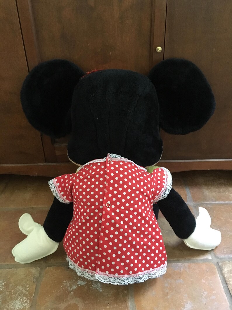 Op de afbeelding: Een Minnie Mouse knuffel, van achteren gezien, met grote zwarte oren en een rode jurk met witte stippen. De jurk heeft witte kanten randen en een kleine knoopsluiting. De armen en handen van de pop zijn zwart en wit.