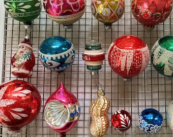 Puede incluir: Una colección de adornos navideños vintage en varias formas y colores, incluyendo rojo, verde, azul y dorado. Algunos adornos presentan diseños intrincados y detalles de purpurina, mientras que otros tienen una forma redonda clásica.