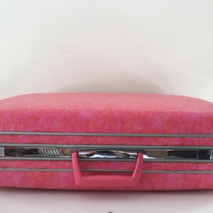 Pu&ograve; includere: Una valigia rigida rettangolare con una finitura testurizzata rosa vivace. Il bagaglio presenta una fascia centrale in metallo color cromo con meccanismi di chiusura integrati e una maniglia in plastica rosa abbinata.