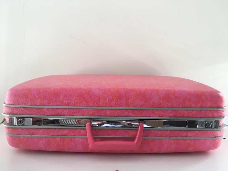Pu&ograve; includere: Una valigia rigida rettangolare con una finitura testurizzata rosa vivace. Il bagaglio presenta una fascia centrale in metallo color cromo con meccanismi di chiusura integrati e una maniglia in plastica rosa abbinata.