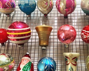 Può includere: Una collezione di decorazioni natalizie vintage in varie forme e colori. Gli ornamenti includono palline rotonde, forme a campana e una figura di Babbo Natale. I colori includono rosa, blu, oro e rosso. Gli ornamenti sono esposti su una griglia metallica.