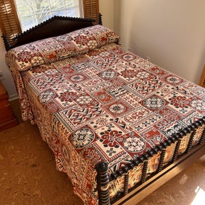 May include: A bed with a dark wooden frame and a patterned quilt. The quilt features a repeating design of red, black, and cream-colored geometric shapes and floral motifs. The headboard is also dark wood with a decorative top.