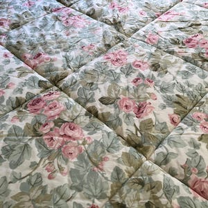May include: A quilted bedspread featuring a repeating floral pattern of pink roses and sage green leaves on an off-white background. The fabric is stitched in a grid pattern, creating a series of soft, padded squares across the entire surface.