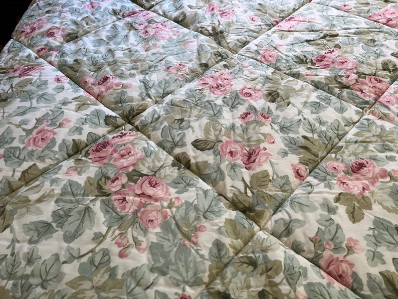 May include: A quilted bedspread featuring a repeating floral pattern of pink roses and sage green leaves on an off-white background. The fabric is stitched in a grid pattern, creating a series of soft, padded squares across the entire surface.