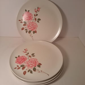 May include: A set of three round off-white ceramic plates featuring a floral pattern of pink roses with green leaves and brown stems. The plates have a smooth surface and a simple design, with the rose motif positioned on the left side of each plate.
