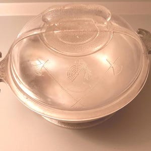 May include: A round metal serving dish with a clear glass lid. The lid features an etched design of a knight's helmet centered between two crossed battle axes. The dish has two textured metal handles on opposite sides for easy lifting and transport.