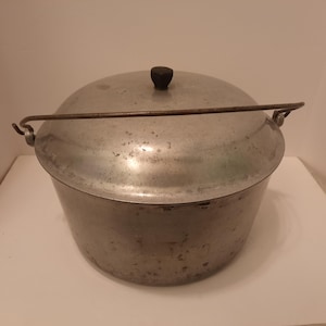 May include: A vintage aluminum cooking pot with a lid and a handle. The pot is round with a slightly flared rim and a black knob on the lid. The handle is a metal bar that arches over the top of the pot.