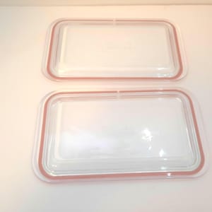 May include: Two clear, rectangular glass lids with a reddish-brown rubber seal. The lids are designed to fit a rectangular container. The image is taken from a top-down perspective, showing the lids against a white background.