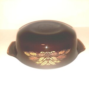May include: A brown Pyrex casserole dish with a wheat pattern in gold and orange. The dish has two handles and a rounded shape. This vintage dish is perfect for baking and serving.