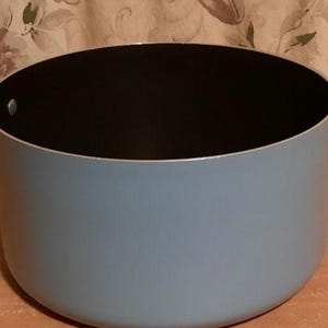 May include: A light blue cooking pot with black interior and two black handles. The pot is round and has a silver rim. The handles are attached to the sides of the pot. The pot is sitting on a wooden surface.