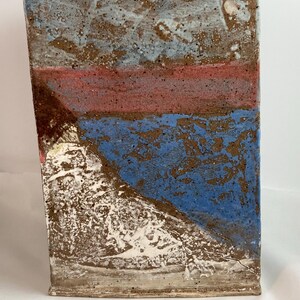 May include: A rectangular ceramic vase with a textured surface. The vase features a color block design with bands of blue, red, and brown. The top section is a mix of blue and brown, the middle is red, and the bottom is blue.