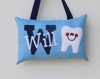 May include: A light blue rectangular pillow with a dark blue ribbon hanger. The pillow features the name Will in dark blue and white embroidered letters, alongside an embroidered white tooth graphic with a smiling face and red cheeks.
