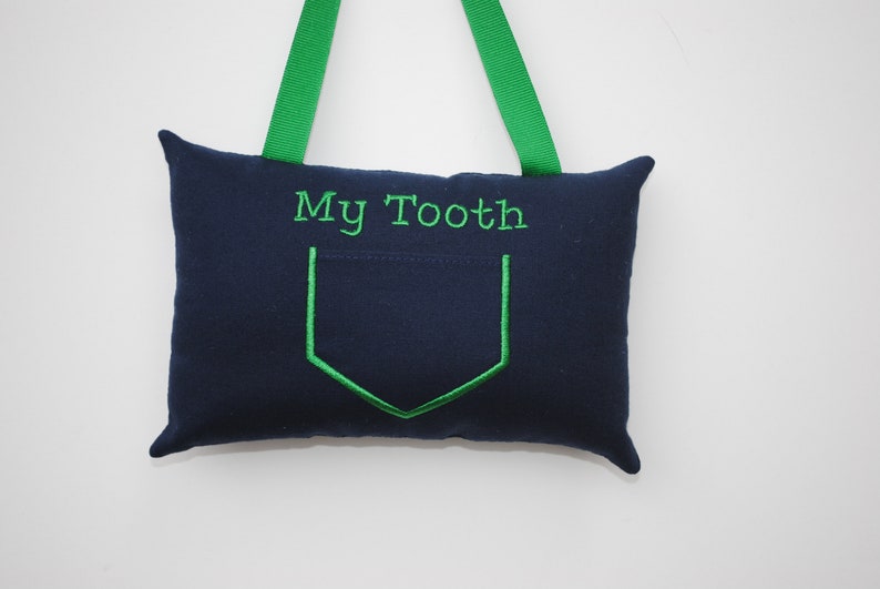 May include: A blue fabric pillow with a green pocket and the text "My Tooth" embroidered on the front. The pillow has green fabric loops for hanging.