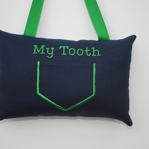May include: A navy blue pillow with a green pocket and the text "My Tooth" embroidered on the front. The pillow has green ribbon loops for hanging.