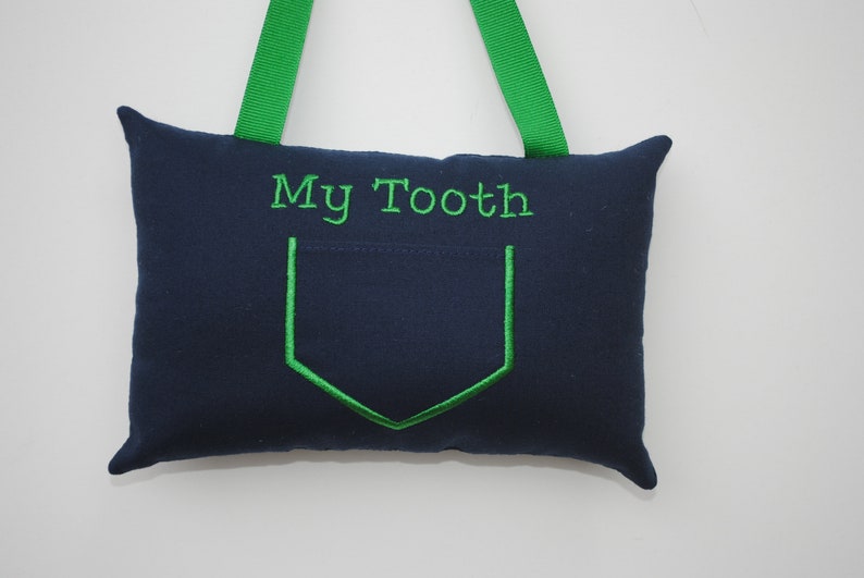 May include: A navy blue pillow with a green pocket and the text "My Tooth" embroidered on the front. The pillow has green ribbon loops for hanging.