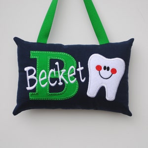 May include: A navy blue pillow with a green embroidered letter "B" and the name "Beckett" in white. A white embroidered tooth with a red smiling face is on the right side of the pillow. The pillow has green ribbon loops for hanging.