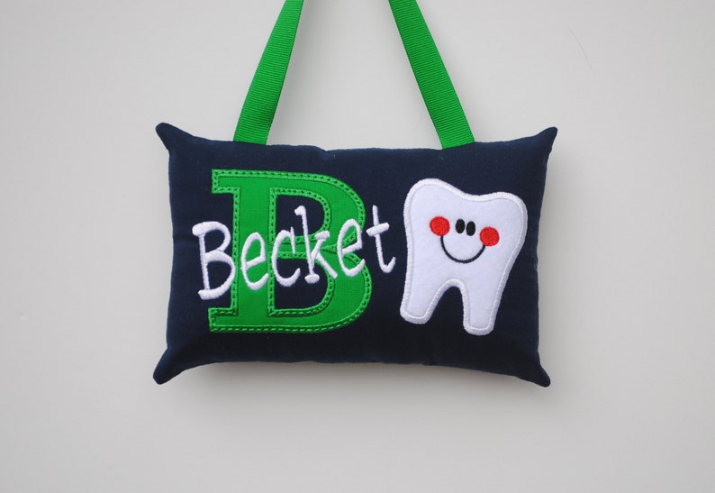 May include: A navy blue pillow with a green embroidered letter "B" and the name "Beckett" in white. A white embroidered tooth with a red smiling face is on the right side of the pillow. The pillow has green ribbon loops for hanging.