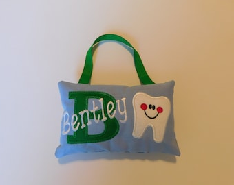 May include: A light blue rectangular pillow with a green ribbon handle. The pillow features an embroidered green letter B, the white stitched name Bentley, and a white tooth applique with a smiling face, pink cheeks, and black eyes.