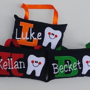 May include: Three personalized tooth pillows with a tooth and initial embroidered on each. The pillows are navy blue with red, orange, and green letters. The names are Kellan, Luke, and Beckett.