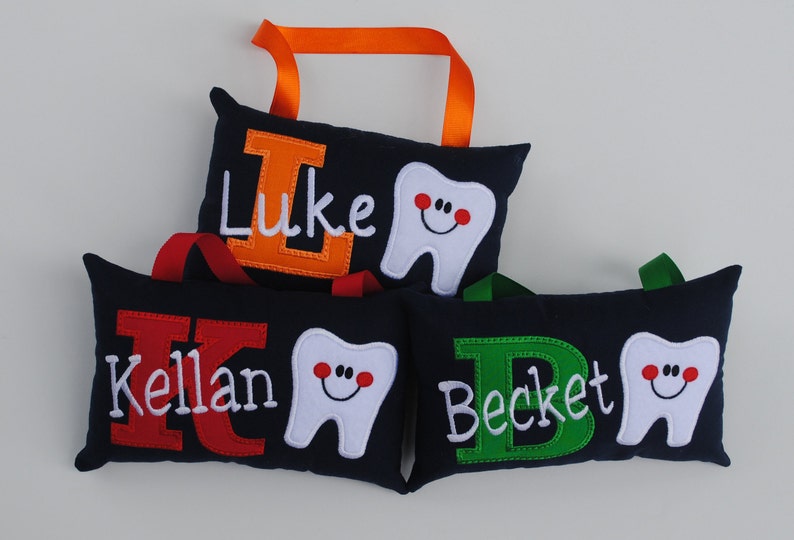 May include: Three personalized tooth pillows with a tooth and initial embroidered on each. The pillows are navy blue with red, orange, and green letters. The names are Kellan, Luke, and Beckett.