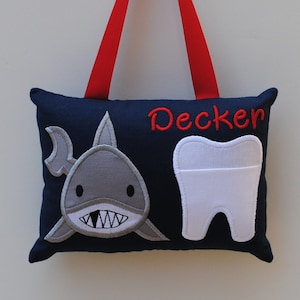 May include: A blue tooth fairy pillow with a shark and a tooth pocket. The pillow has a red ribbon for hanging and the name "Decker" embroidered on it.