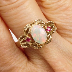 May include: A gold ring featuring an oval opal center stone with iridescent colors. Two small, round, red gemstones flank the opal. The ring has an ornate, textured gold filigree design.