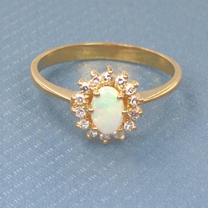 May include: A gold ring featuring a central oval opal gemstone surrounded by a halo of small, clear gemstones. The ring has a simple, polished gold band. The ring is set against a light blue textured background.