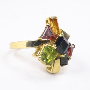 Op de afbeelding: Gouden ring met een cluster van groene, rode en zwarte edelstenen. De ring heeft een eenvoudige band en de stenen zijn in een geometrisch patroon gezet.