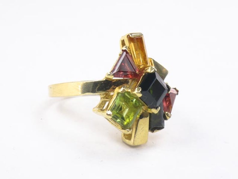 Op de afbeelding: Gouden ring met een cluster van groene, rode en zwarte edelstenen. De ring heeft een eenvoudige band en de stenen zijn in een geometrisch patroon gezet.